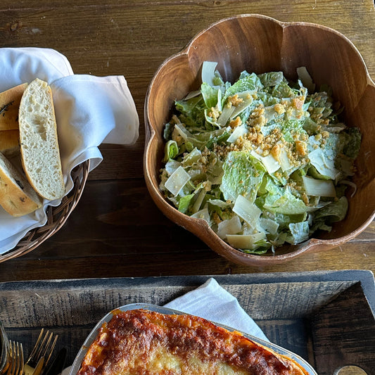 Wooden tray with lasagna, salad, bread, and cutlery on a wooden surface
