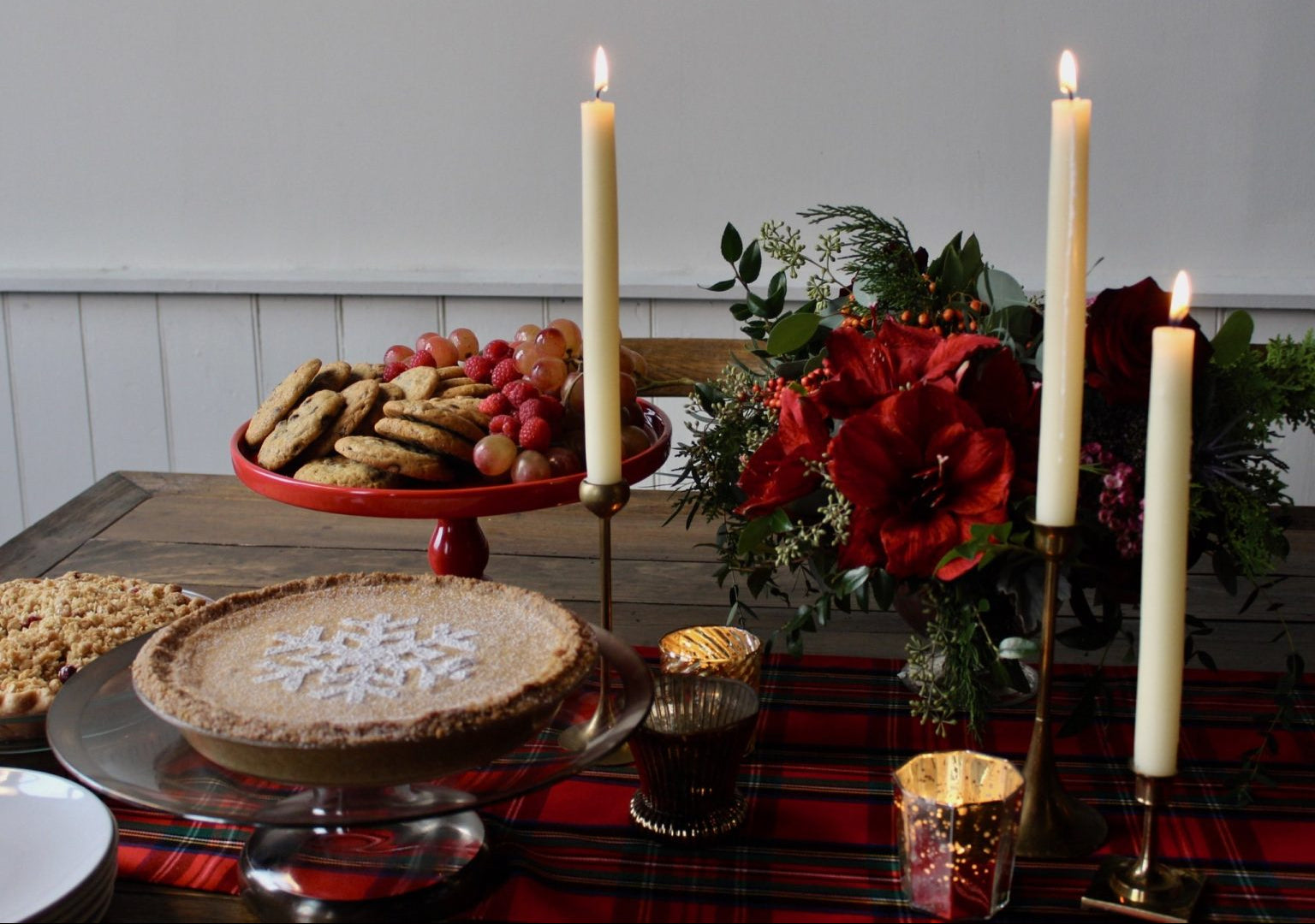 Dining table set with a pie, cookies, fruits, and candles on a plaid tablecloth.